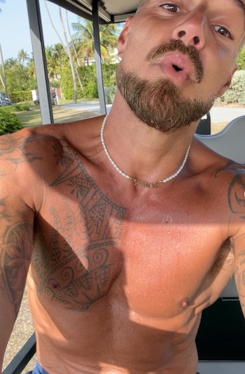 Hombre sin camiseta en traje de baño saltando en la arena de una playa, con el mar, rocas, árboles y cielo nublado