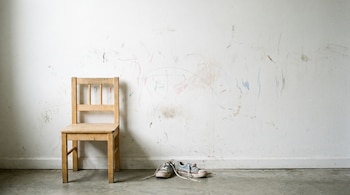 Silla de madera pequeña desgastada y par de zapatillas infantiles desatadas sobre suelo gris, junto a pared blanca con dibujos de crayón.