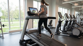 Una mujer trota sobre una cinta de correr en un gimnasio junto a máquinas elípticas y grandes ventanales con vista al exterior.