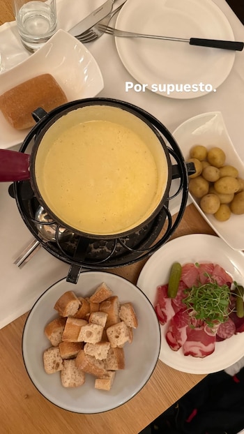 Vista aérea de una mesa con una olla de fondue de queso humeante, rodeada de platos con patatas cocidas, pan en cubos, y embutidos con encurtidos