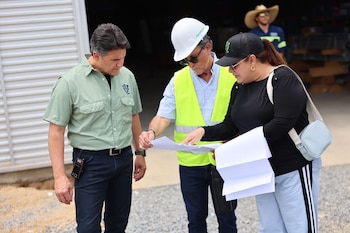 Tres personas, dos hombres y una mujer, revisan planos al aire libre. Un hombre lleva camisa verde, otro chaleco reflectante y casco blanco. La mujer usa gorra negra