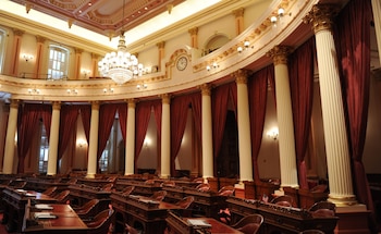 Un hemiciclo legislativo vacío con filas de escritorios de madera, columnas blancas y cortinas rojas debajo de un balcón y un candelabro