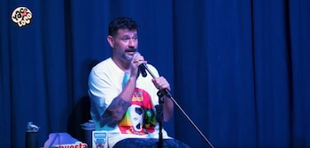 Yaco Eskenazi, con barba y camiseta blanca con estampado, sostiene un micrófono en un escenario con telón azul, botellas y cajas en primer plano