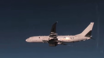 Un avión militar de fuselaje estrecho, de color gris claro con marcas "NAVY" en el costado, vuela horizontalmente contra un cielo azul oscuro