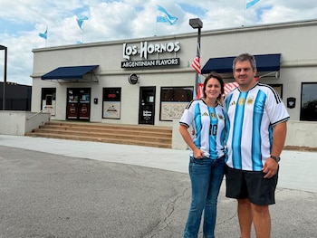 Una pareja de pie frente al Restaurante Los Hornos. Ambos visten camisetas de fútbol de Argentina. Banderas argentinas y una estadounidense ondean sobre el edificio