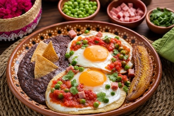 Plato de barro con dos huevos fritos sobre tortillas con salsa, guisantes, jamón, frijoles negros con totopos y una pieza de plátano frito.