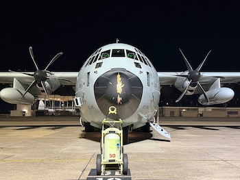 Parte frontal de un avión militar gris iluminado de noche en una pista de aterrizaje, con las escaleras de la puerta abiertas