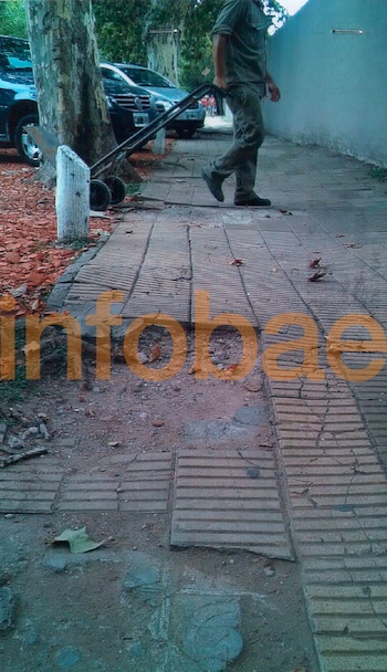 Vereda deteriorada con baldosas rotas y tierra, un tronco de árbol en el lateral y la rueda de un automóvil, con el logo de Infobae