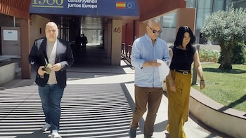 Tres personas caminan por una acera soleada frente a un edificio moderno con la bandera de España y la Unión Europea. Dos hombres y una mujer