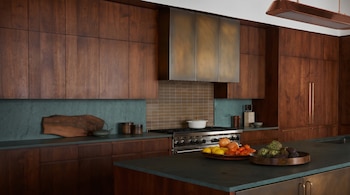 Una cocina moderna con gabinetes de madera oscura, encimeras verdes, una estufa de acero inoxidable, campana de bronce, y una isla con frutas y verduras.