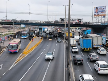 Autopista con tráfico de vehículos, autobuses y camiones. Un puente vehicular elevado. En la calzada hay una motocicleta y una persona tendida. Muro con grafitis y cartel publicitario