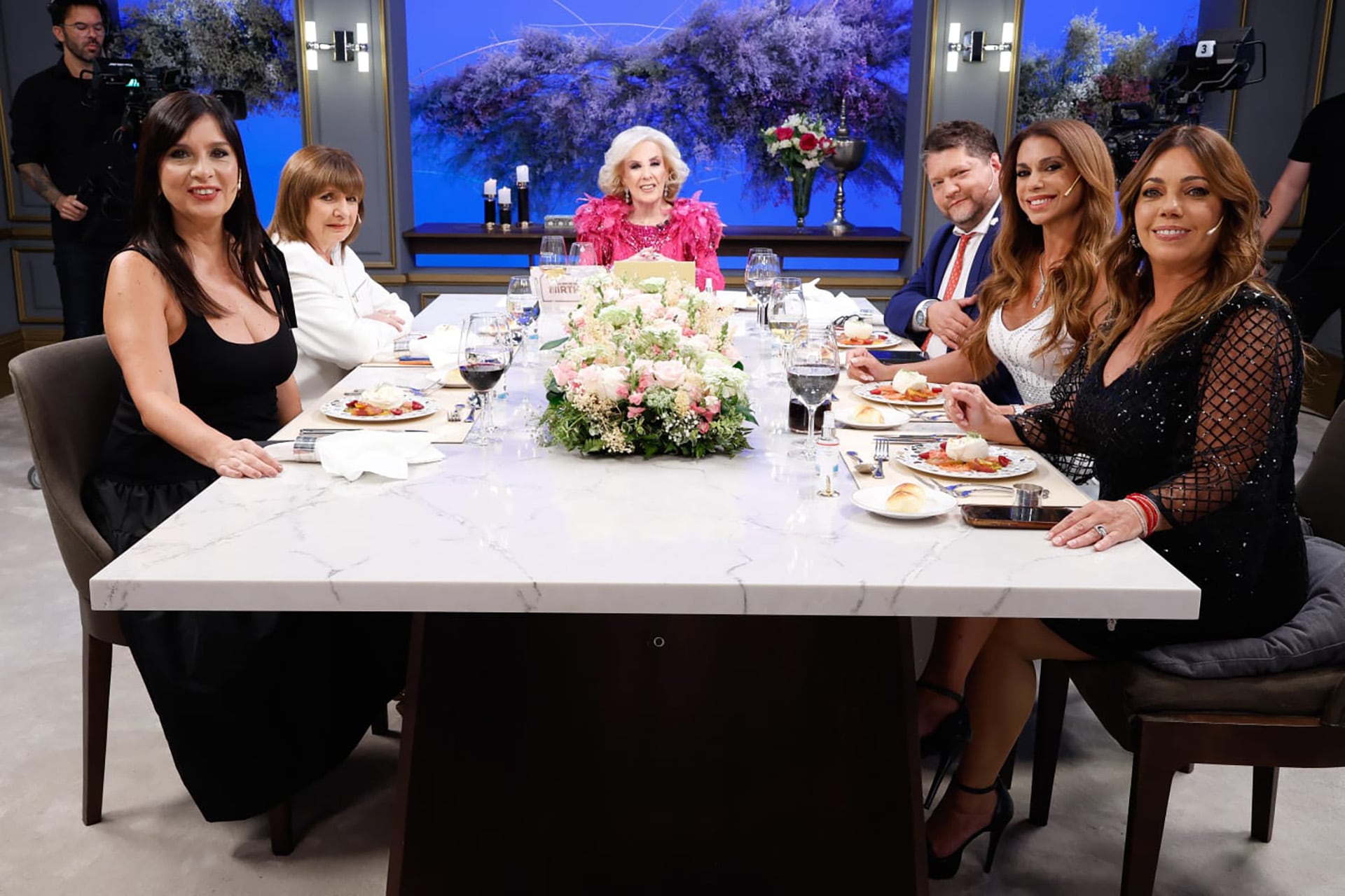 Mirtha Legrand, vestida de un vibrante color rosa, preside una elegante mesa con un centro floral, compartiendo un momento con sus invitados en el set de su programa televisivo (Imagen Ilustrativa Infobae)