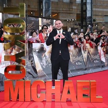 Un hombre con traje negro hace la forma de un corazón con sus manos detrás de grandes letras doradas y rojas que dicen Tokyo y Michael