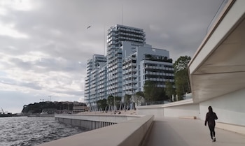 Un edificio moderno de gran altura sobre la costa junto a un paseo marítimo de hormigón donde camina una persona frente al mar