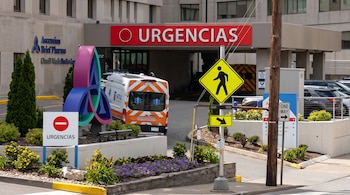 Entrada de urgencias de un hospital con un cartel rojo, una ambulancia blanca y naranja y varias señales de tránsito.