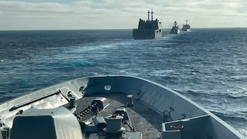 Vista desde la proa de un buque de guerra gris, mostrando otros tres buques militares grises navegando en fila sobre el mar azul bajo un cielo nublado
