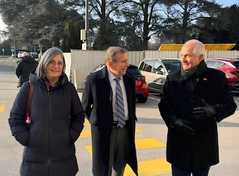Tres personas adultas, dos hombres y una mujer, de pie en un exterior con árboles y vehículos de fondo. El suelo tiene líneas amarillas