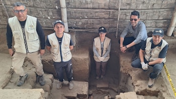 Cinco personas con chalecos institucionales posan alrededor de una trinchera de excavación arqueológica bajo una techumbre de caña