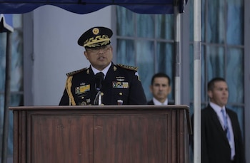 Oficial del ejército con uniforme de gala azul marino y medallas, hablando en un atril de madera. Dos hombres de traje desenfocados en el fondo