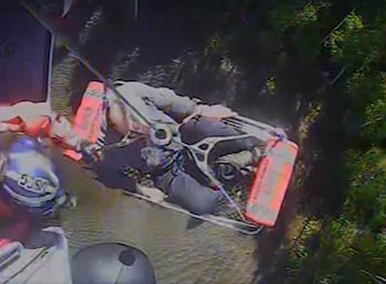 Una camilla de rescate suspendida por un cable sobre vegetación y agua junto a un rescatista con casco visto desde la altura