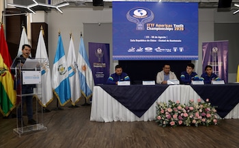 Hombre en podio, cinco banderas, banner grande azul, mesa con mantel, cuatro personas sentadas, carteles de nombre, arreglo floral, banner vertical morado