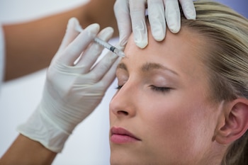 Una mujer rubia con ojos cerrados recibe una inyección en la frente. Dos manos enguantadas sostienen la jeringa y la frente