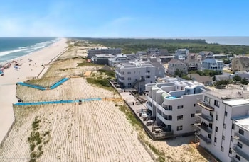 Vista aérea de una larga playa de arena dorada con gente y sombrillas, dunas, y edificios residenciales a lo largo de la costa bajo un cielo azul claro