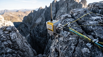Dispositivo metálico de monitoreo sísmico con antena y cables fijado a una roca de montaña con picos al fondo.