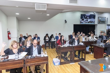 Vista aérea de una sala de reuniones donde varias personas, sentadas en escritorios de madera, tienen las manos levantadas votando. Una pantalla muestra una videollamada