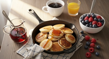 Mini pancakes con jarabe en una sartén, junto a bayas frescas, café, jugo de naranja y un jarro de jarabe, todo sobre una mesa de madera.