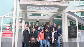 Un grupo de dieciocho personas sonriendo, de pie frente a la entrada de un edificio moderno de la Universidad de Lodz, Polonia, con letreros en polaco