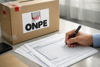 Detalle de una mano rellenando un acta electoral con un bolígrafo, junto a una urna de cartón de la ONPE con precintos, sobre una mesa gris.