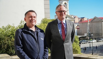 Dos hombres de pie al aire libre: uno viste una chaqueta azul oscura y el otro un traje gris con corbata roja y anteojos