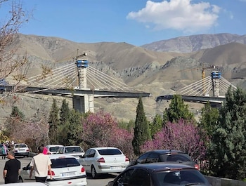 Puente B1 en construcción con dos torres de hormigón y cables tensores blancos. Grúas en las torres. Montañas áridas, cielo azul y coches con personas en primer plano