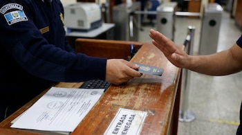 Manos de oficial de Migración de Guatemala sosteniendo pasaporte, frente a una mano levantada. Letrero "ENTRADA DENEGADA" en mostrador.