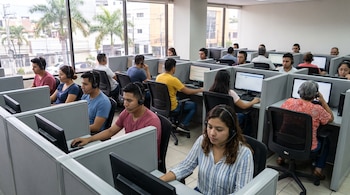 Varios empleados sentados en módulos de oficina usan computadoras de escritorio y auriculares frente a monitores en una sala iluminada.