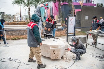 Varias personas manipulan una gran pieza de piedra con herramientas manuales y eléctricas. Cables, postes y palmeras al fondo, con una carpa y fardos de heno