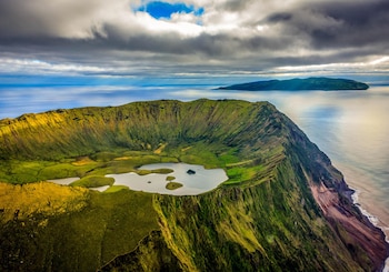 Cráter volcánico con una laguna interior y laderas verdes junto al mar, bajo un cielo nublado con otra isla visible al fondo