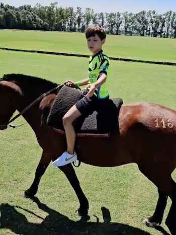Un niño con camiseta deportiva verde fluorescente y pantalones cortos negros monta un caballo marrón sobre césped