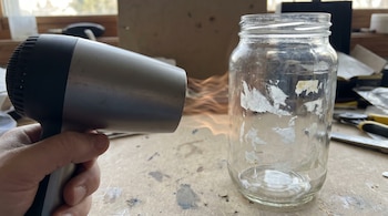 Primer plano de una mano sujetando un secador de pelo plateado y negro, que emite aire caliente hacia un frasco de vidrio transparente con residuos de pegatinas.