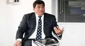 Hombre sentado con traje oscuro, camisa blanca y corbata de puntos. Tiene un tablet y documentos sobre su regazo. Se observan persianas en el fondo