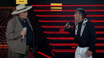 Un hombre con sombrero de ala ancha y otro con chaqueta decorada sostienen micrófonos en un escenario con luces rojas.