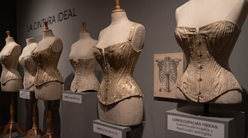 Cinco corsés antiguos en maniquíes, con un diagrama de un esqueleto torácico y letreros informativos en una exposición.