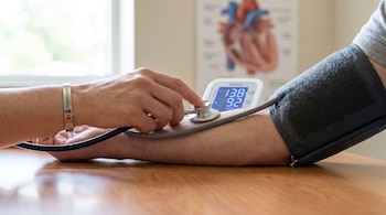 Brazo adulto con manguito de presión arterial sobre mesa de madera. Una mano sostiene un estetoscopio. Monitor digital indica 138/92 mmHg. Brazalete médico, ilustración de corazón.
