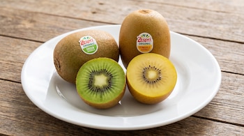 Cuatro kiwis, dos enteros y dos cortados por la mitad, se encuentran en un plato blanco sobre una mesa de madera. Los kiwis son de variedades verde y dorada.