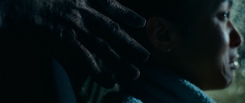 Una mano oscura y arrugada se acerca al perfil de un rostro humano, apenas iluminado, con un fondo borroso de luces en la oscuridad