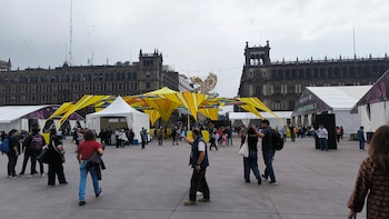 Peatones caminan en una plaza de explanada gris rodeada por edificios históricos, carpas blancas y una estructura decorativa amarilla
