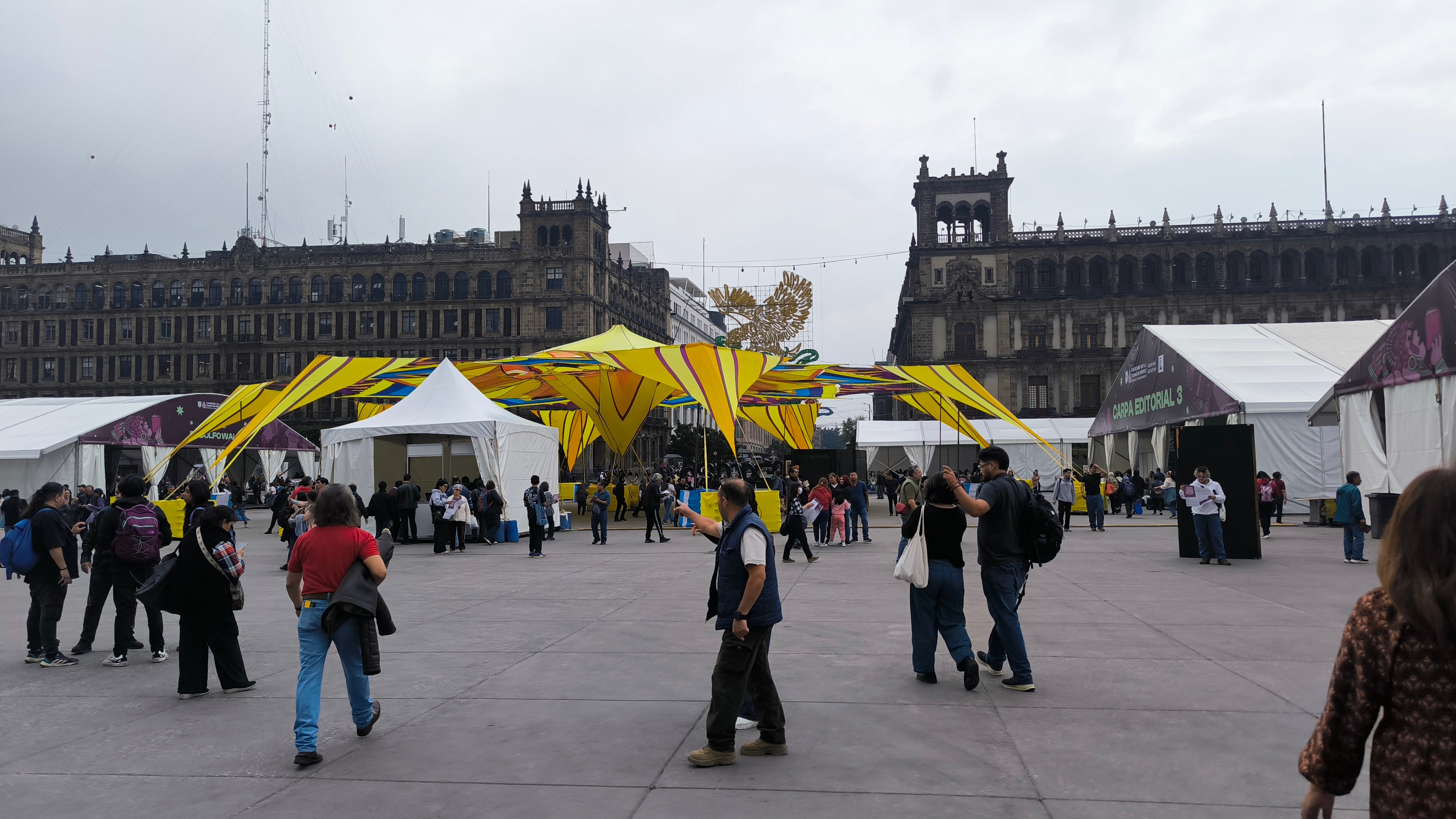Varias personas transitan por la plaza del Zócalo frente a carpas editoriales y estructuras amarillas instaladas para una feria al aire libre. (Selene Miranda/Infobae México)
