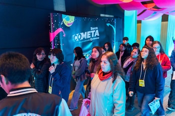 Un grupo diverso de personas, incluyendo mujeres y jóvenes, caminan o se detienen en un espacio con luces azules y un cartel de "Beca Cometa"