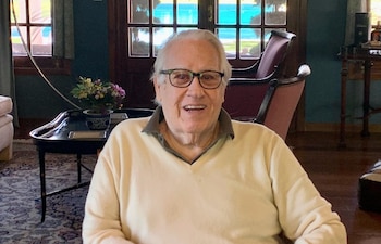 Un hombre de edad avanzada con gafas y suéter beige sonríe en una habitación con sillones, mesas de madera y un ventanal